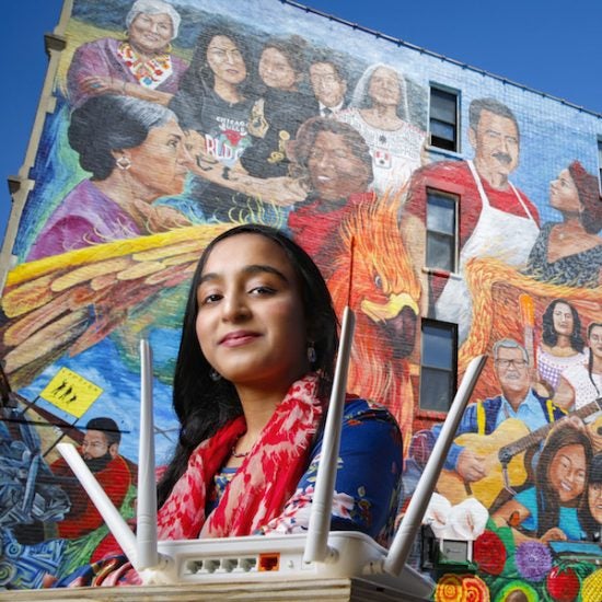 a female student poses in between a colorful Chicago community mural in Pilsen and a router that will help expand internet access in that community